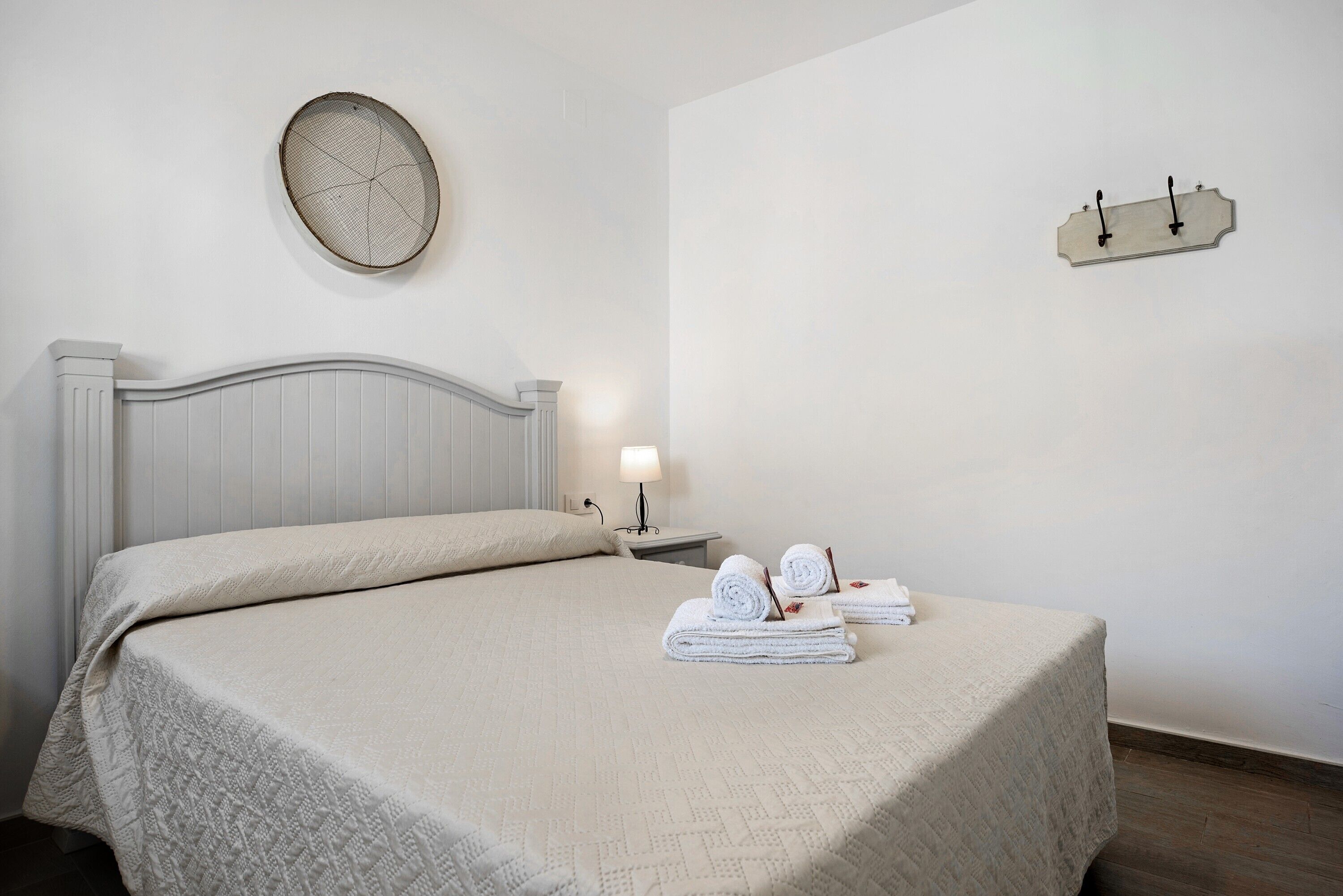A neatly made bed with a textured bedspread, a round wall clock, a bedside table with a lamp, and a wall-mounted towel rack with towels.