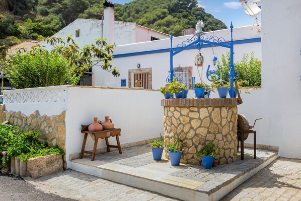 A courtyard with stone walls, potted plants, a stone well, and a wooden table with pottery.