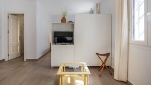 Un espacio habitable pequeño con una televisión en un estante, una mesita, una silla y una ventana con cortinas.