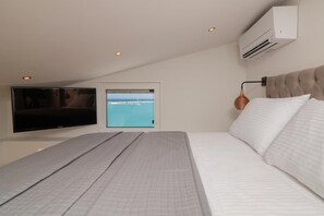 A bedroom with a bed, a television, an air conditioning unit, and a window with a view of the sea.