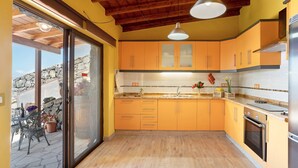 Een keuken met houten vloer, oranje kastjes, een roestvrijstalen koelkast en een stenen muur zichtbaar door een schuifdeur.