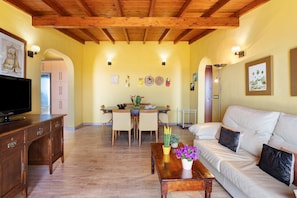 Un salon avec un plafond en bois, un canapé, une table basse décorée de fleurs, un téléviseur posé sur un meuble en bois, et un coin repas avec une table et des chaises.
