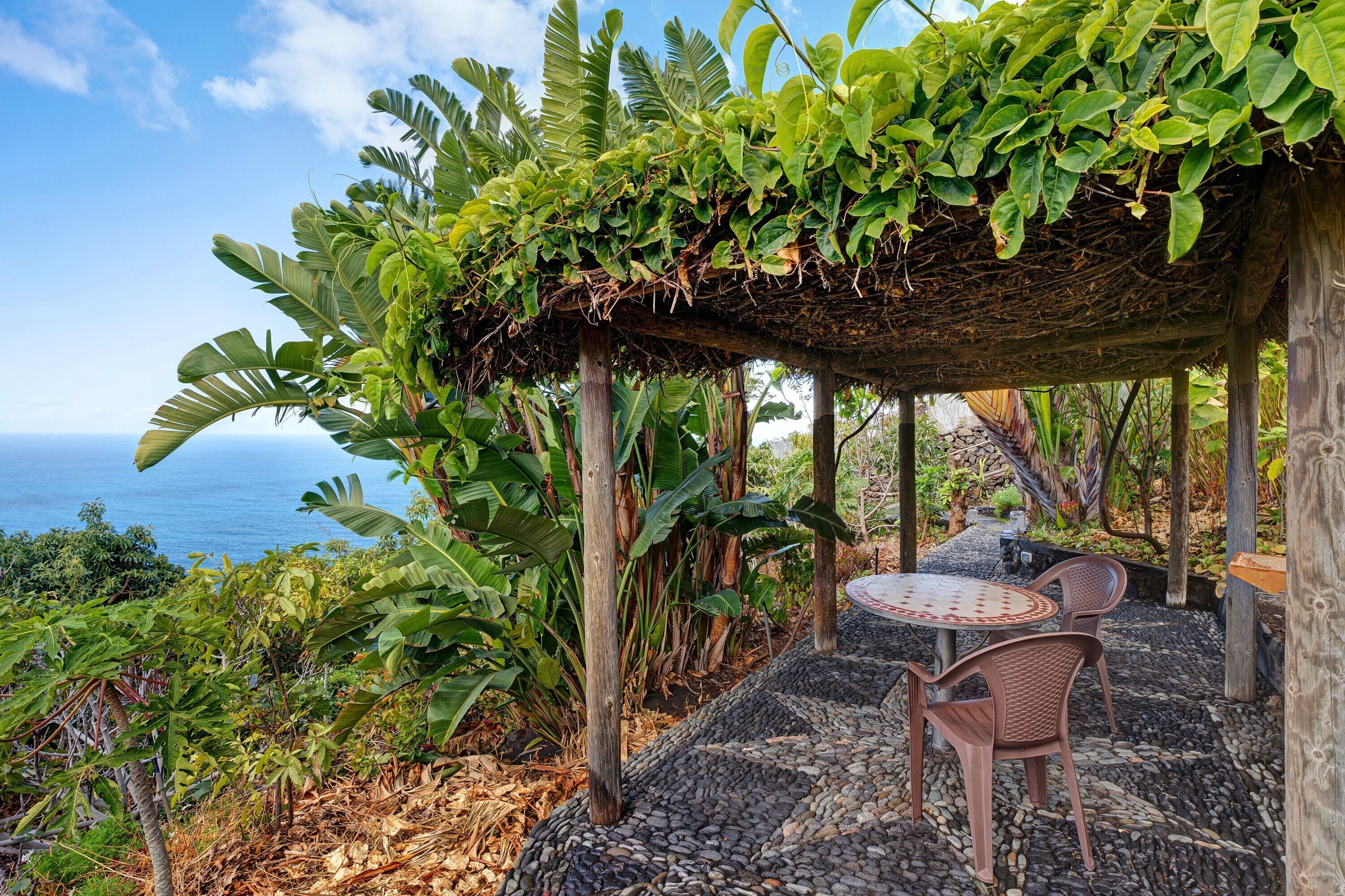 A covered outdoor seating area with a table and chairs, surrounded by lush greenery and overlooking the ocean.