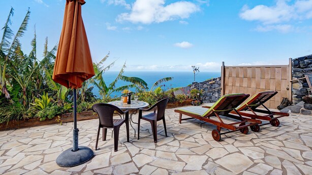 Een terras met een tafel, stoelen, een parasol en lounge fauteuils, met uitzicht op de oceaan.