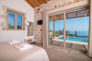 A bedroom with a large bed, a TV, a stone wall, and a view of a pool and ocean. - Spartia Suites - Brand New Seaview Suites - Spartia Suites - Brand New Seaview Suites (Zakynthos)