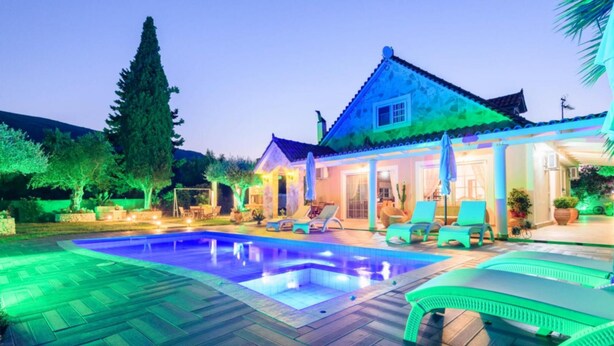 A swimming pool with a tiled deck, a covered patio area with lounge chairs, and a house with a tiled roof and columns. - Zante Sun I - Getaway Villa - Zante Sun I - Getaway Villa (Zakynthos)