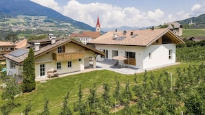 A residential building with a balcony, a small garden, and a view of a mountain and church in the background.