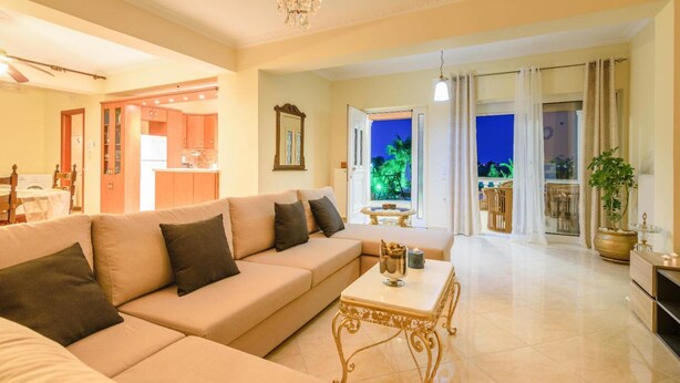 A spacious living room with a beige sofa, a dining table, and a chandelier. There is a kitchen area visible in the background, and a balcony with a view of the night sky.