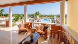 A balcony with wicker furniture, a glass table, and a view of a swimming pool and greenery.
