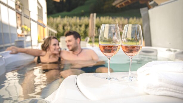 Ein Whirlpool mit zwei Gläsern Roséwein, einem weißen Handtuch und Blick auf ein Gebäude und Grünflächen.