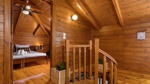 Een houten blokhut van binnen met een slaapkamer op de zolder, een trap en een plafondventilator.