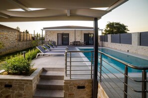 A pool area with a stone wall, a covered seating area, and a building with a tiled roof. - Lea Villa - Brand New Modern Villa - Lea Villa - Brand New Modern Villa (Zakynthos)