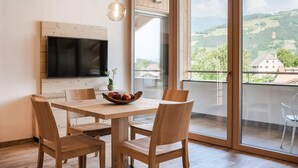 Un coin repas comprenant une table, des chaises, un saladier de fruits, un téléviseur et une grande fenêtre donnant sur un paysage.