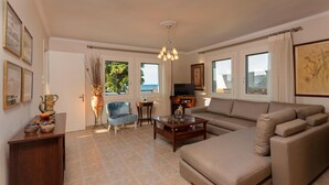 A living room with a sofa, a chair, a coffee table, and a TV. There are windows with curtains, a vase with branches, and framed pictures on the walls. - Villa Elizabeth - Seafront Modern Villa - Villa Elizabeth - Seafront Classic Villa (Zakynthos)