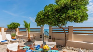 An outdoor dining area with a wooden table set for two, surrounded by white cushioned chairs, a large tree, and a view of the sea. - Villa Elizabeth - Seafront Modern Villa - Villa Elizabeth - Seafront Classic Villa (Zakynthos)
