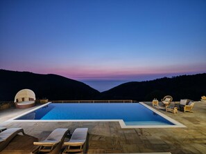 A swimming pool with lounge chairs and a dome structure, surrounded by hills and a sunset sky.