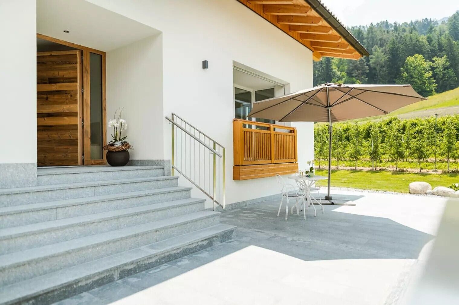 A modern house with a wooden door, a staircase, a patio with a table and chairs, and a view of a green landscape.