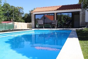 Ein Schwimmbecken mit klarer, blauer Wasserlinie, eine überdachte Terrasse mit Stühlen und ein Steinhaus mit einem Ziegeldach.