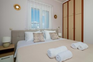 A neatly made bed with white linens and two rolled towels, a window with sheer curtains, a wooden wardrobe, and a small round wall decoration.