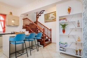 A modern kitchen with a wooden staircase, blue bar stools, and a bookshelf with decorative items.