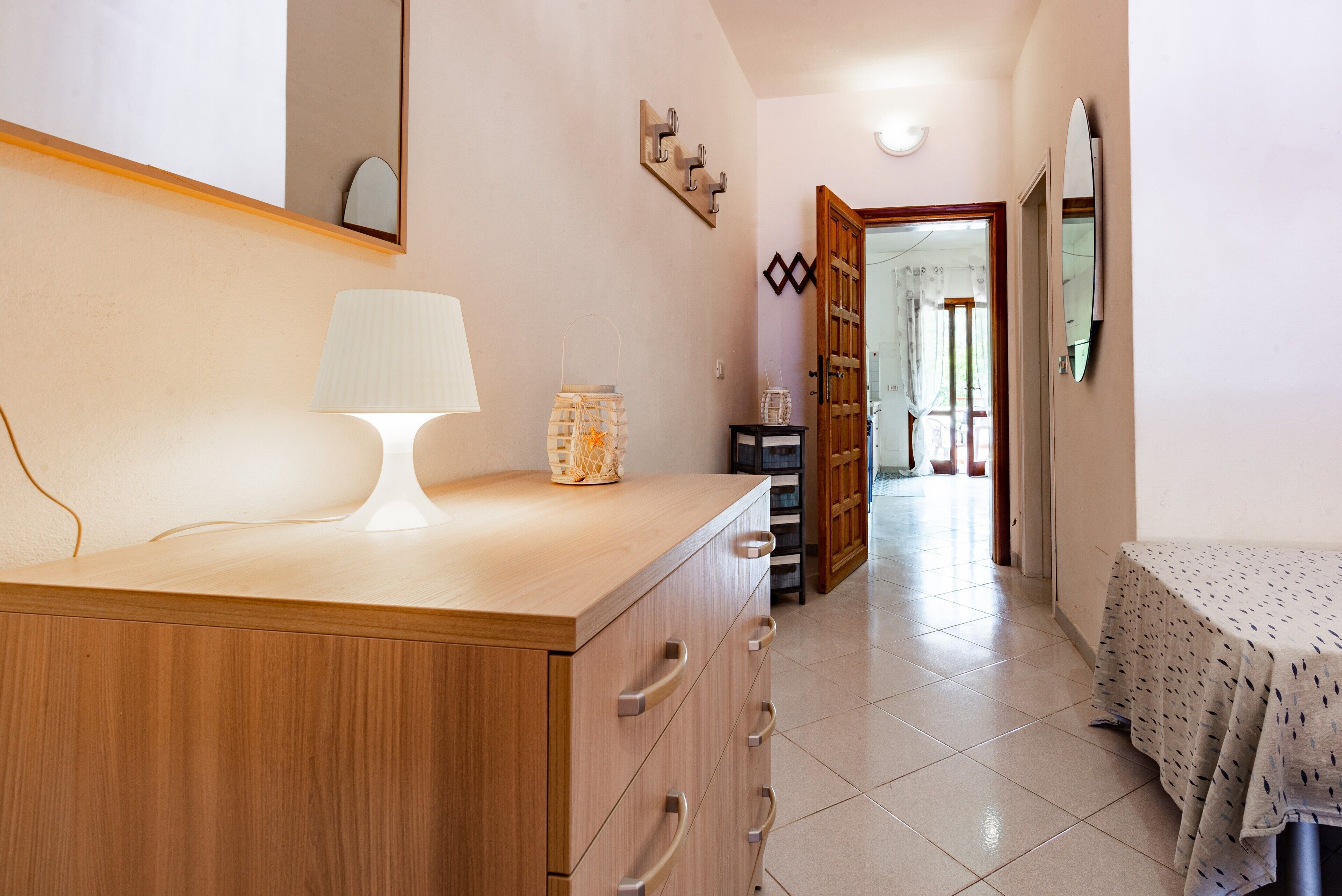 A bedroom with a bed, a bedside table with a lamp, a dresser, and a door leading to another room.