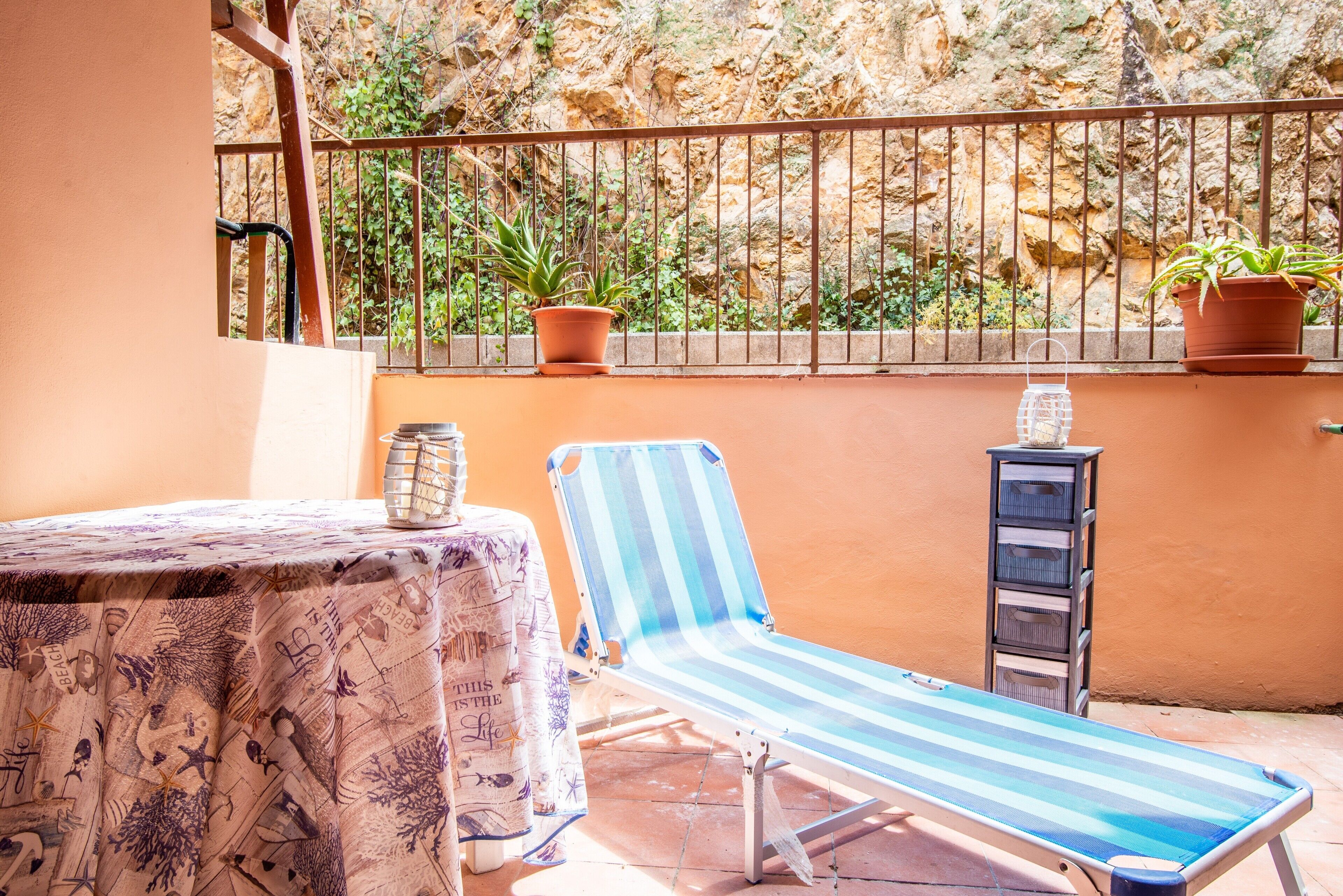 A balcony with a striped lounge chair, a table with a patterned tablecloth, potted plants, and a metal storage unit.