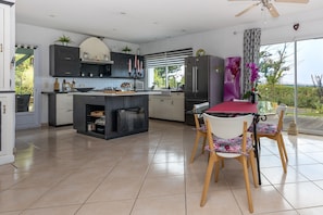 A modern kitchen with a central island, a dining area with a red table, and a view through large windows.
