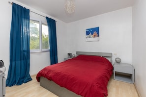 Quarto com cama, mesa de cabeceira, abajur, quadro na parede e janela com cortinas.