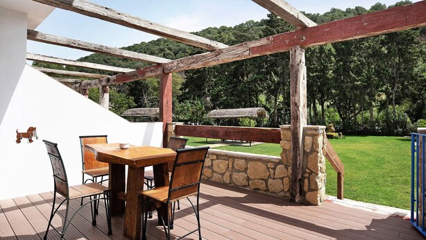 Um pátio externo com mesa e cadeiras de madeira, uma parede de pedra e vista para uma paisagem verde.