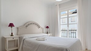 A white bed with a headboard, two bedside tables with lamps, a window with a view of buildings, and a balcony. - Holiday Home 'Casa Begines' close to City Centre with Shared Pool (Vejer)