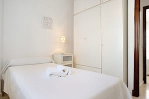 Un lit bien fait, avec une serviette pliée, une lampe de chevet et une armoire, dans une chambre propre et blanche.