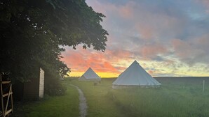 Un terrain de camping avec deux tentes blanches et une cabane en bois, entouré d’herbe et d’arbres, au coucher du soleil.