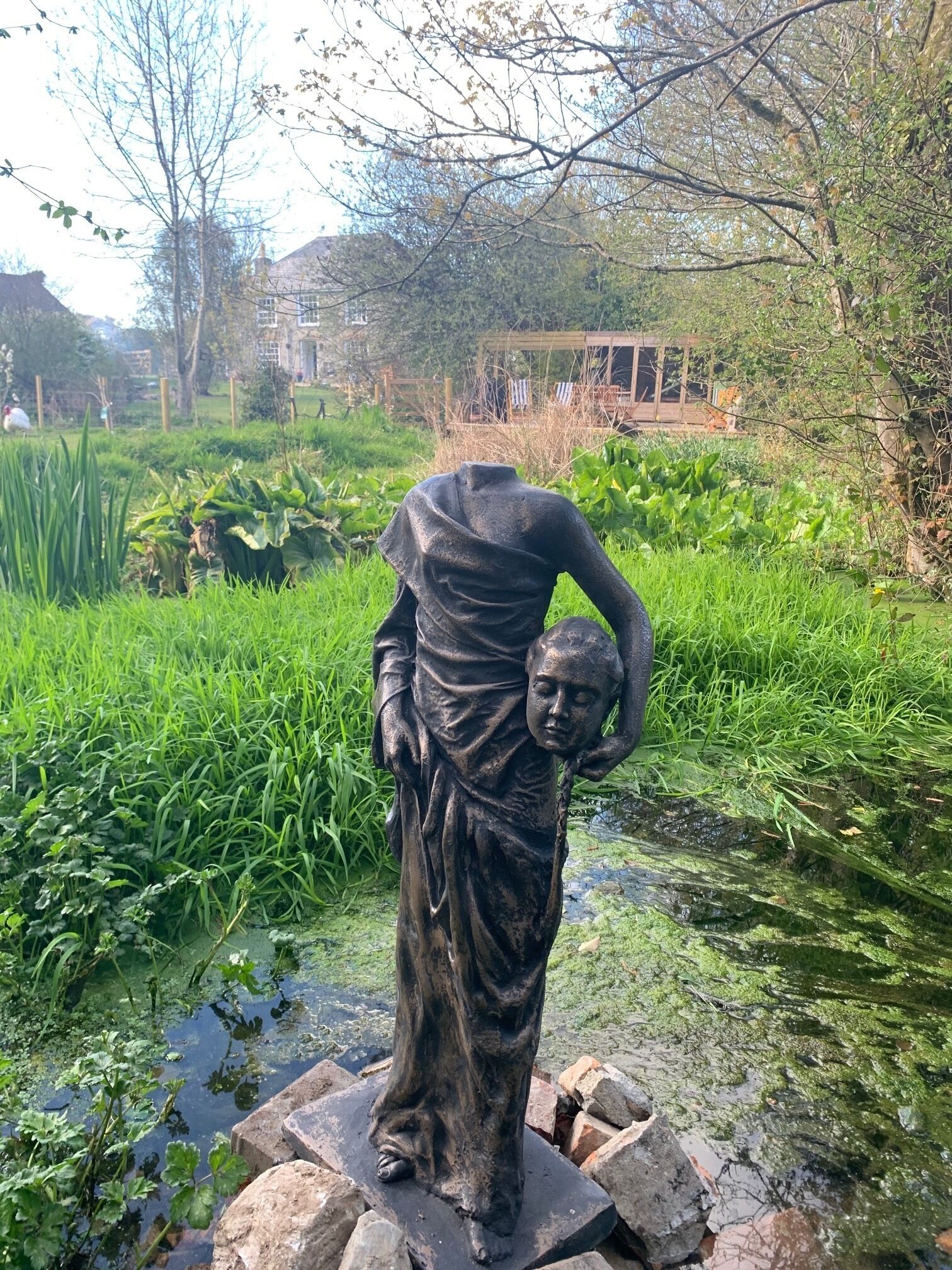 A statue in a garden with a pond, surrounded by greenery and a wooden structure in the background.