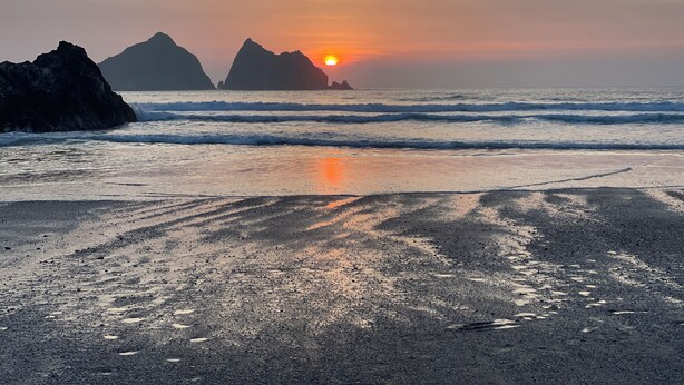Une plage avec des vagues, un coucher de soleil et des formations rocheuses à l’horizon.