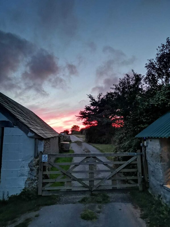 Una caseta rural con una puerta cerrada, rodeada de vegetación y un atardecer de fondo.