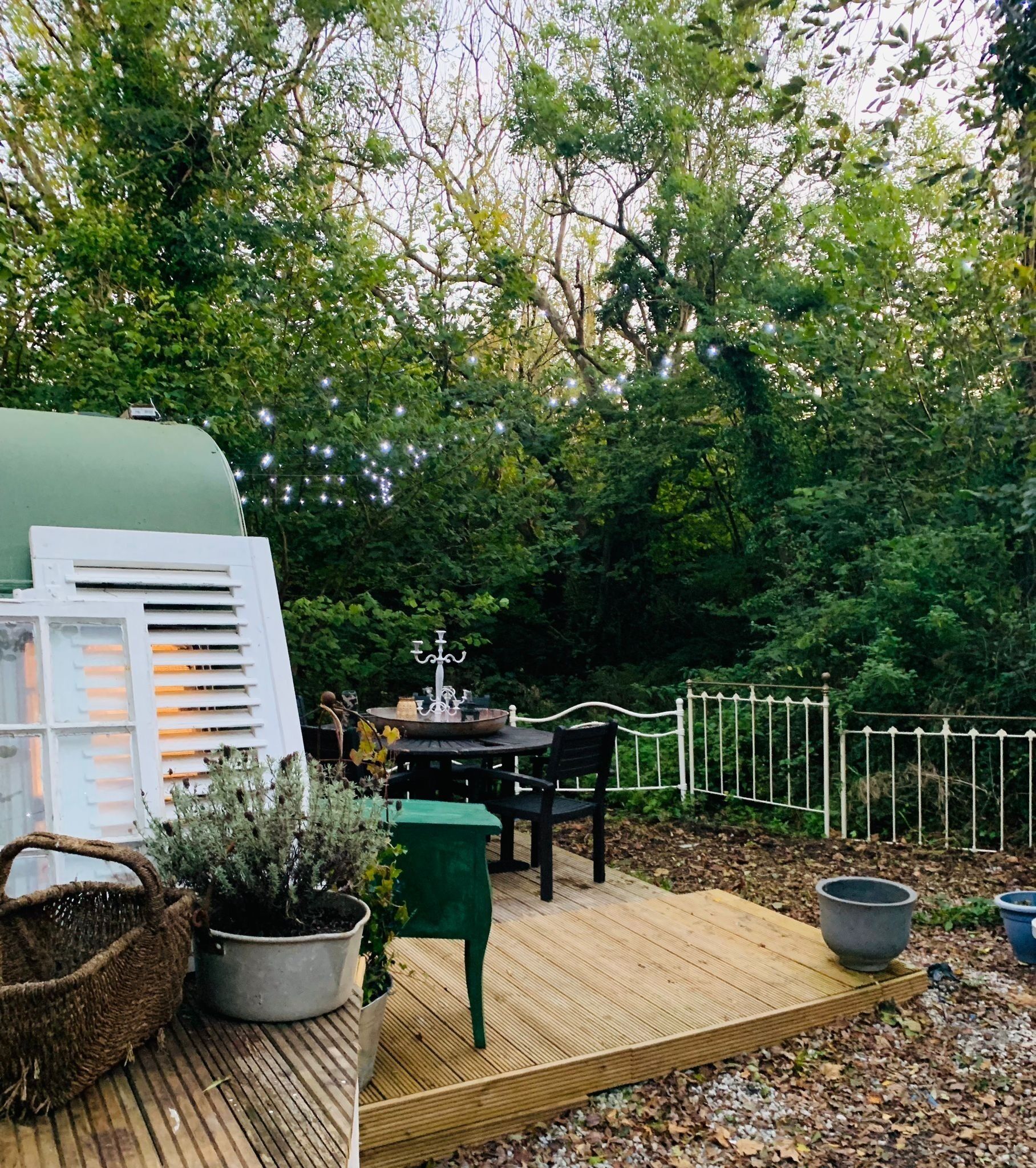 A wooden deck with a table and chairs, surrounded by greenery and a white fence.