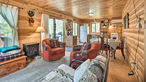 A cozy cabin interior with a fireplace, wooden walls, a dining area, and a living room with a sofa and chairs.