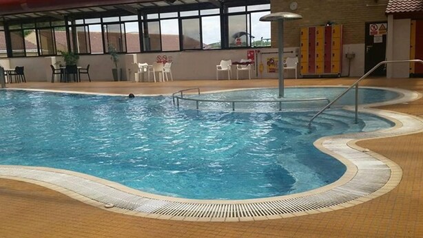 Una piscina interior amb sòl de rajola i zona de vora piscina amb cadires i taules.