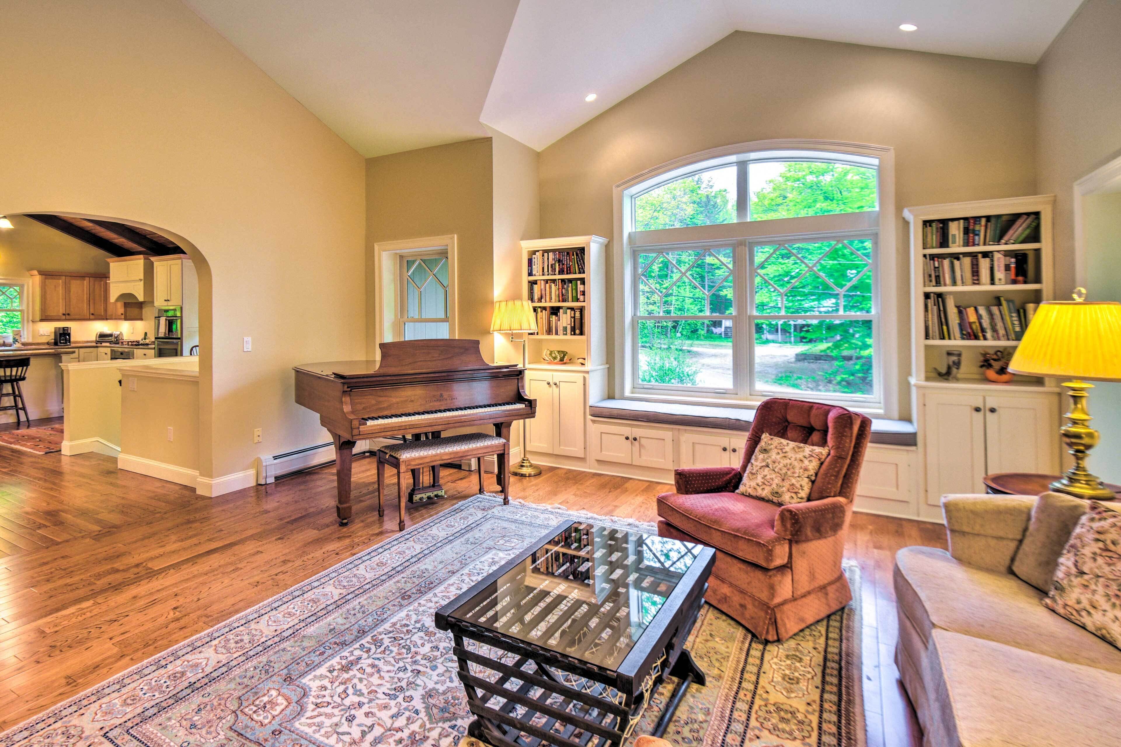 A spacious living room with a grand piano, a red armchair, a glass coffee table, and a large window offering a view of the outdoors.
