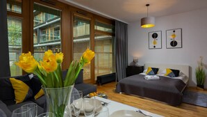Una habitación de hotel moderna con una cama grande, un sofá, una mesa de comedor preparada para dos personas y un jarrón con flores amarillas.
