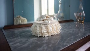 Une table ronde recouverte d’une nappe en dentelle, avec un objet blanc posé dessus, un miroir reflétant une fenêtre et une bouteille, et un plan de travail en marbre.