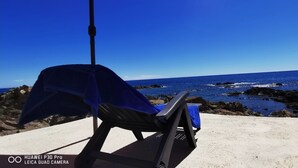 A beach chair with a blue towel and umbrella, facing a rocky coastline and clear blue sea.