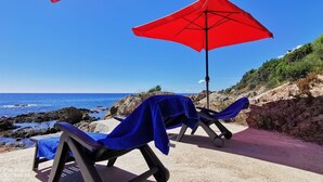 Two beach chairs with blue towels and a red umbrella on a rocky seaside path.