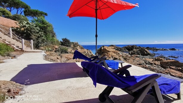 A beachside lounge with two sunbeds, a red umbrella, and a rocky coastline.