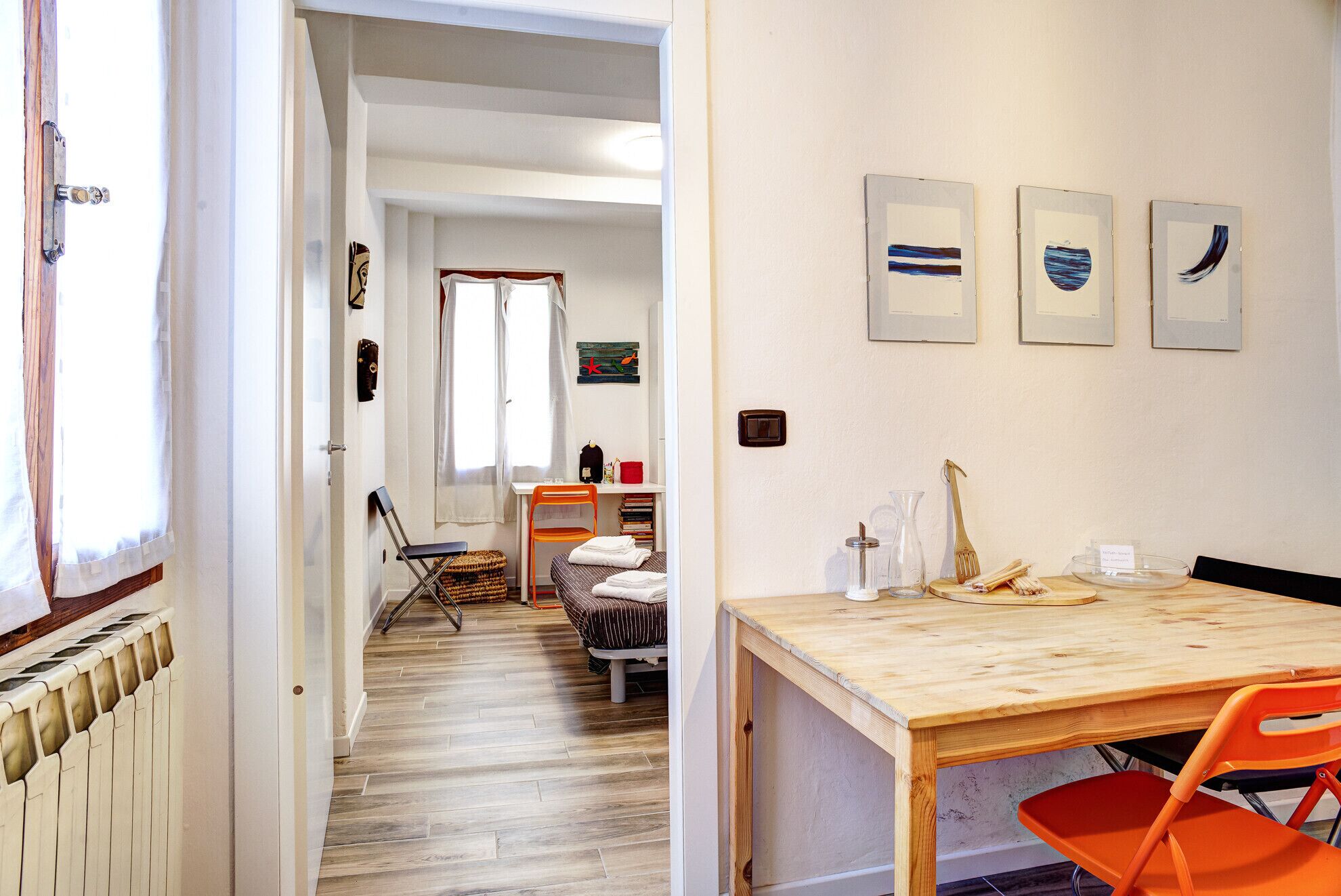 Ein Zimmer mit Holzboden, einem Esstisch mit Stühlen, einem Heizkörper, einem Fenster mit Vorhängen und Wandbildern.