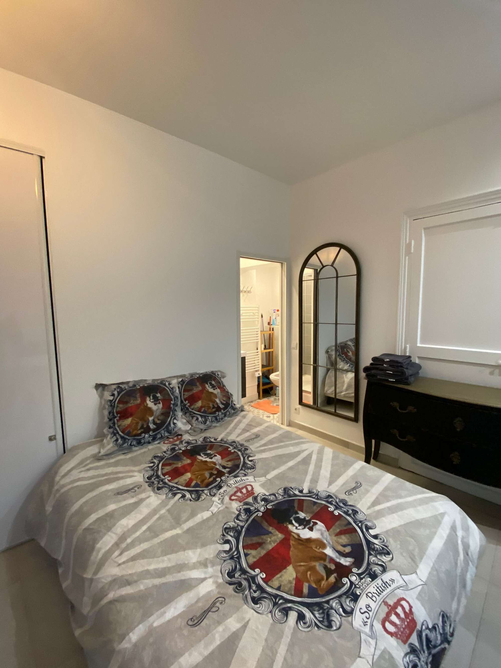 A bedroom with a bed featuring a patterned bedspread and decorative pillows, a dresser with a mirror, and a view into an adjacent room.