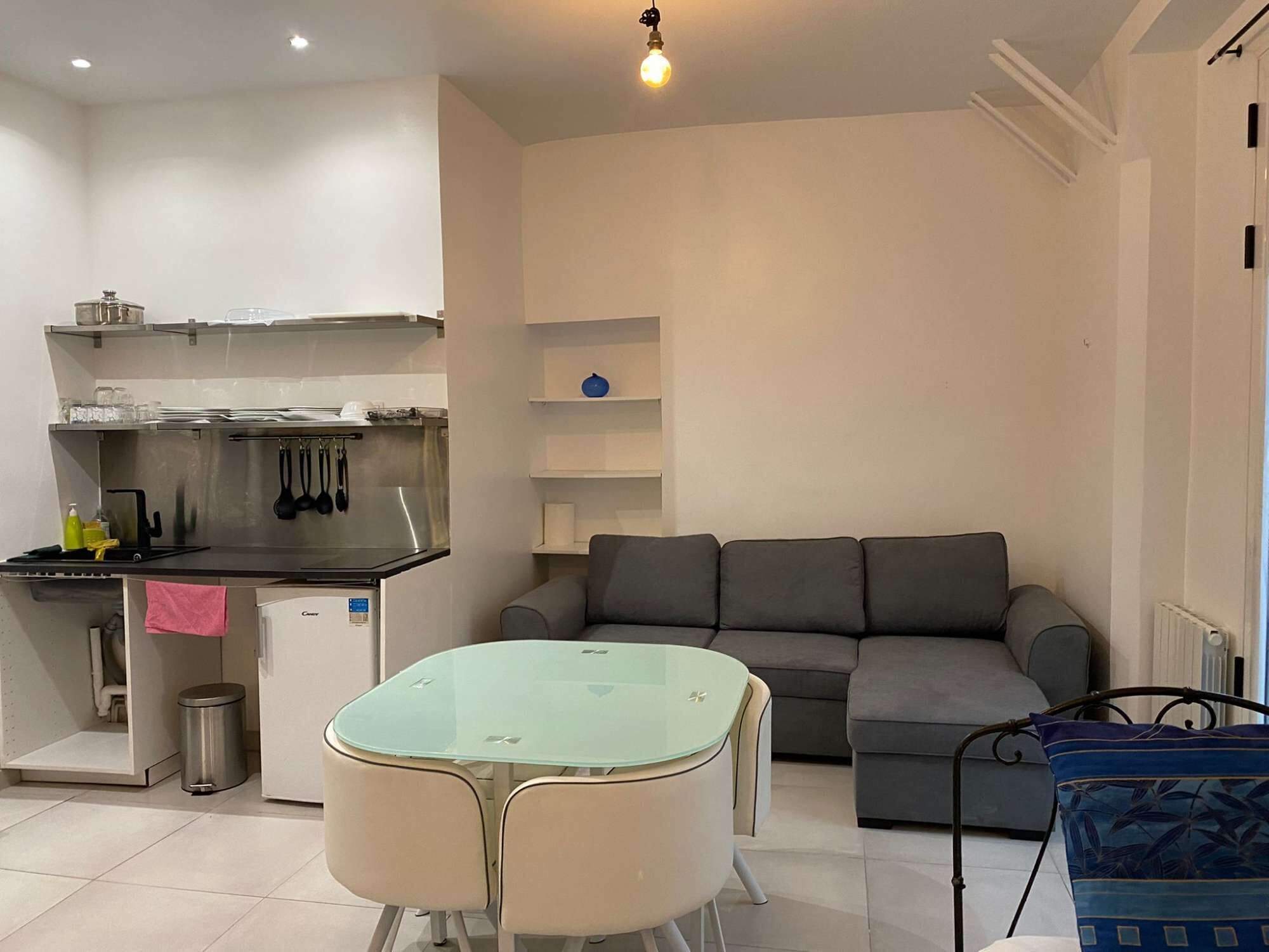 A compact living space with a kitchenette, a glass-top dining table, a grey sofa, and a patterned cushion.
