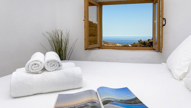 A bedroom with a bed, two rolled white towels, a window with a view of the sea, and a magazine on the bed. - Country house 15 minutes from the beach (Málaga)