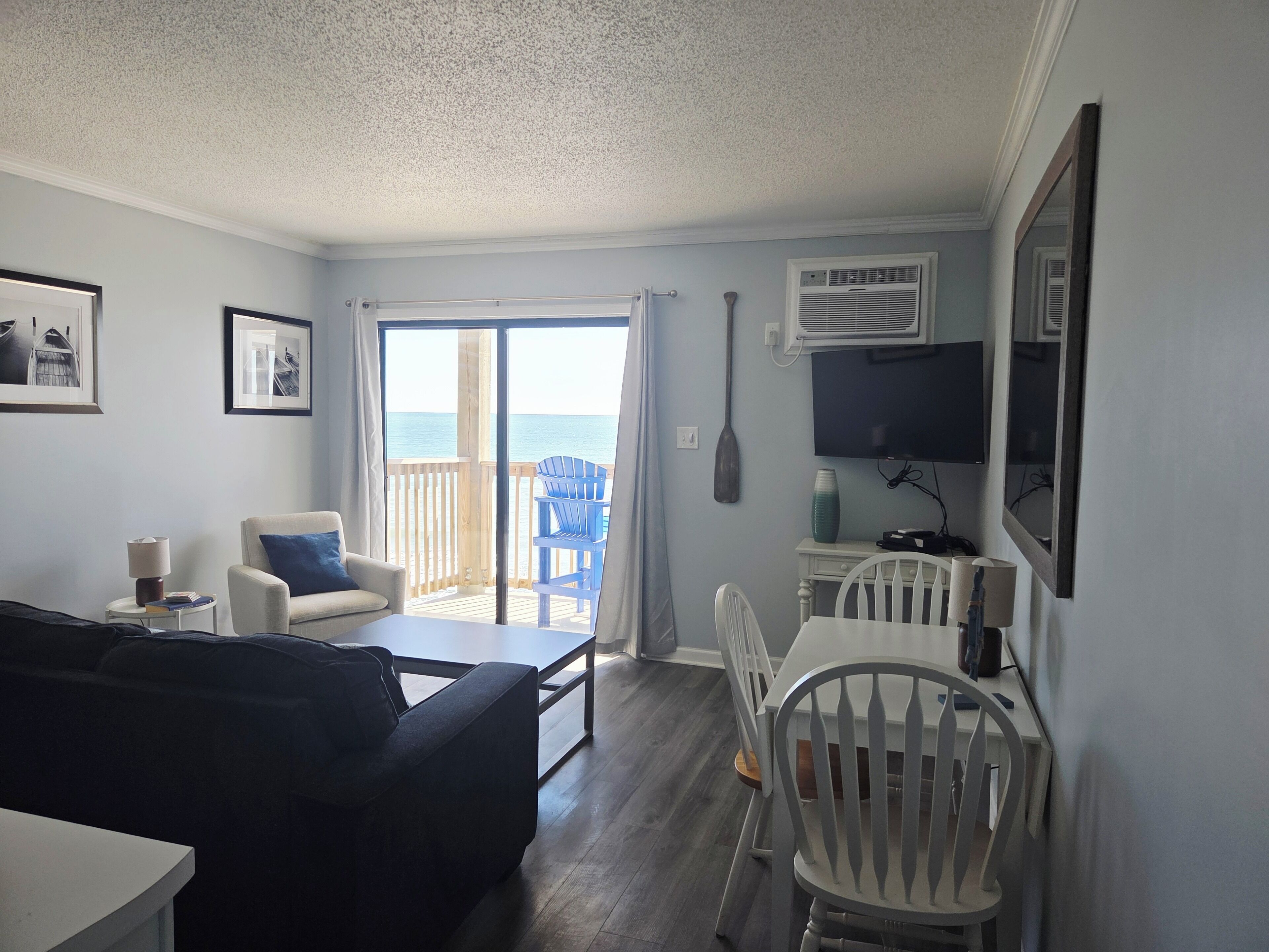 A room with a sofa, a dining table with chairs, a TV, and a view of the ocean through a sliding door.