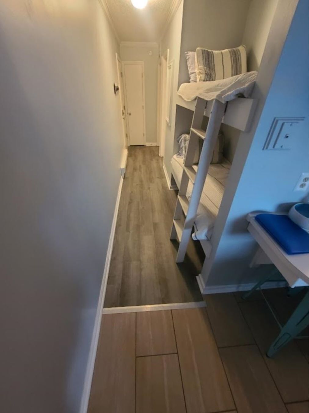 Ein schmaler Flur mit Holzboden, einer weißen Tür am Ende und einem Etagenbett mit blauem Kissen auf der rechten Seite.